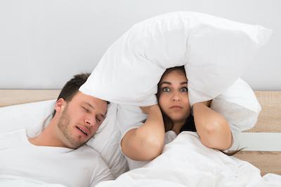 woman holding a pillow covering her ears while her husband snores loudly next to her
