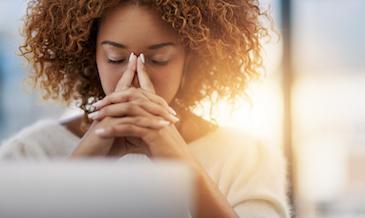  A woman sits at her laptop, covering her face with her hands while her eyes are closed