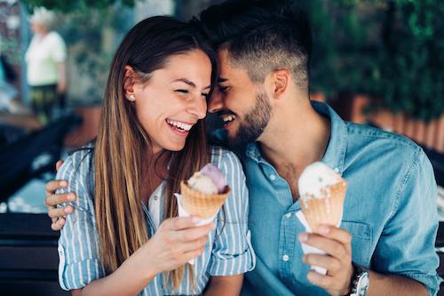 A man and woman sit on a bench, enjoying ice cream cones together on a sunny day in troy